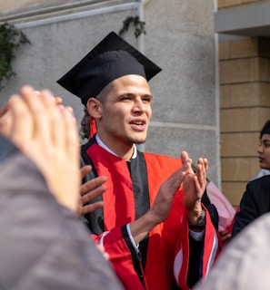 A graduate wearing a red and black gown and cap is clapping, surrounded by people. The setting appears to be outdoors near a building.