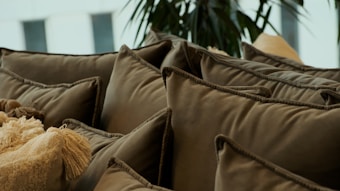 A collection of brown cushions with tassels piled together, creating a cozy and inviting setting. The background showcases some indoor plants, adding a touch of greenery.