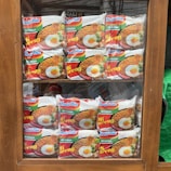 Packets of instant noodles are neatly arranged in a wooden display cabinet. The packaging features the brand 'Indomie Mi Goreng' with an image of cooked noodles topped with a fried egg. The display is organized in two rows, making the products clearly visible through the glass.