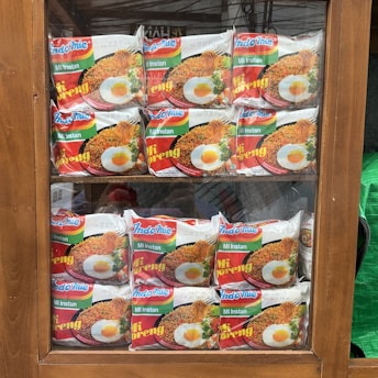Packets of instant noodles are neatly arranged in a wooden display cabinet. The packaging features the brand 'Indomie Mi Goreng' with an image of cooked noodles topped with a fried egg. The display is organized in two rows, making the products clearly visible through the glass.