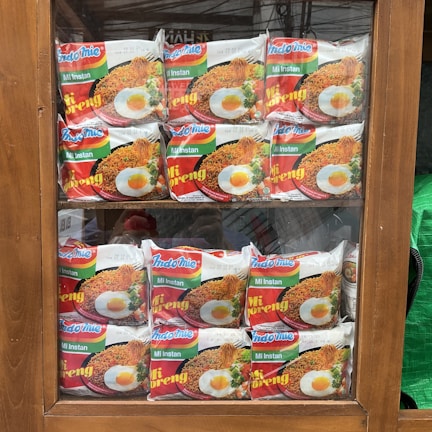 Packets of instant noodles are neatly arranged in a wooden display cabinet. The packaging features the brand 'Indomie Mi Goreng' with an image of cooked noodles topped with a fried egg. The display is organized in two rows, making the products clearly visible through the glass.