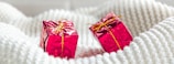Two small, pink gift boxes wrapped in shiny foil with golden ribbons are placed on a soft, textured, white fabric.
