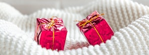 Two small, pink gift boxes wrapped in shiny foil with golden ribbons are placed on a soft, textured, white fabric.