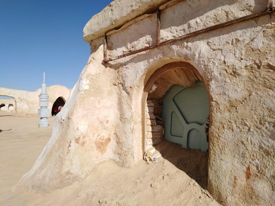 An arid, desert environment with a sandy-colored, textured structure resembling an ancient or sci-fi building. The structure has an archway entrance with a pale blue door featuring geometric patterns. In the background, additional structures and a cylindrical object with vertical sections are visible under a clear blue sky.