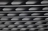 A close-up view of an architectural ceiling featuring repetitive geometric patterns and linear lighting. The design consists of overlapping dark panels with bright strips of light in between, creating a modern and structured appearance.