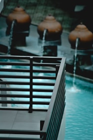 The image contains a decorative outdoor water feature with several spherical terracotta pots, each streaming water into a pool. The water is a striking turquoise, contrasting with the dark metal railing in the foreground.
