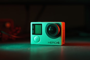 A compact action camera sits on a flat surface, bathed in contrasting red and green lighting. The lens protrudes slightly, and the body features an LCD screen and a prominent power button. The model name 'HERO4' is visible on the front.