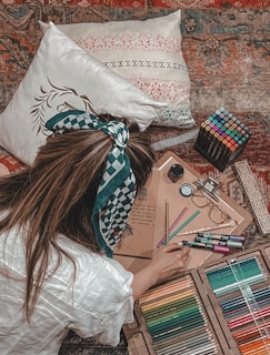 A person is sitting on a patterned carpet surrounded by art supplies. The individual has long hair tied with a patterned band and wears a white shirt. Around them are colored pencils, markers, a sketchbook, glasses, and a ruler. Two decorative pillows are visible in the background.