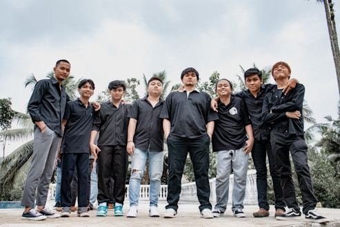 A group of eight young men stand together outdoors, wearing mostly black clothing. Some are smiling while others have neutral expressions. They are surrounded by palm trees and a cloudy sky in the background.