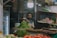 A vendor stands at a market stall sorting green chilies. The setting includes various vegetables like tomatoes, ginger, and cauliflower surrounding the stall. Overhead, a bright light illuminates the area, highlighting the busy atmosphere of the market. Behind the vendor, shelves are stocked with bags and other products, and signage in a local language is visible.