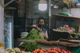 A vendor stands at a market stall sorting green chilies. The setting includes various vegetables like tomatoes, ginger, and cauliflower surrounding the stall. Overhead, a bright light illuminates the area, highlighting the busy atmosphere of the market. Behind the vendor, shelves are stocked with bags and other products, and signage in a local language is visible.