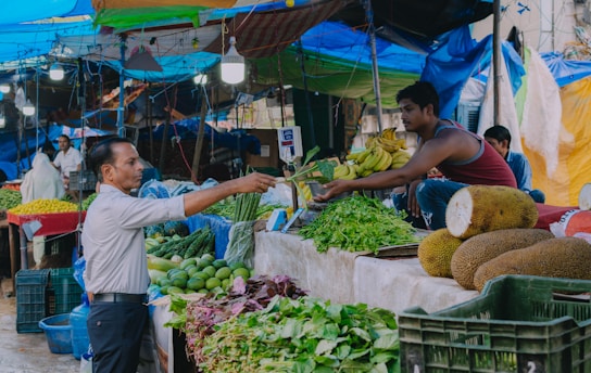 A man in a market is handing money to a vendor who is selling a variety of fresh vegetables and fruits. The vendor is sitting on a raised platform with an assortment of produce including green leafy vegetables, bananas, jackfruit, and other items. The market is covered with colorful tarpaulins and is well-lit with hanging lights. Other people can be seen in the background attending to their stalls.