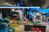 A bustling marketplace scene with various vegetables displayed on stalls. Fresh produce like cucumbers, tomatoes, squashes, and other greens are arranged neatly. Several people can be seen in the market, one man is standing and another is busy organizing vegetables. The market is covered with vibrant blue and black tarps providing shade.