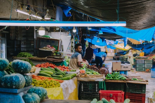 A bustling marketplace scene with various vegetables displayed on stalls. Fresh produce like cucumbers, tomatoes, squashes, and other greens are arranged neatly. Several people can be seen in the market, one man is standing and another is busy organizing vegetables. The market is covered with vibrant blue and black tarps providing shade.