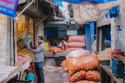 A busy market scene with a man handling a crate, surrounded by sacks of potatoes and onions. The market stall is filled with produce, and another person is visible in the background. The setting includes rustic elements such as worn-out walls and tarpaulin sheets above.