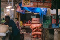A market stall with colorful sacks of produce, particularly onions and potatoes, stacked high with a person sitting behind the counter. Crates and a variety of goods are scattered around the area, and there are signs with Hindi text hanging above. The setting appears to be a bustling market environment.