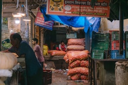 A market stall with colorful sacks of produce, particularly onions and potatoes, stacked high with a person sitting behind the counter. Crates and a variety of goods are scattered around the area, and there are signs with Hindi text hanging above. The setting appears to be a bustling market environment.