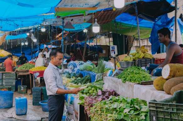 A bustling market scene with a variety of fresh produce on display, including bananas, greens, and jackfruits. Several people are engaged in buying and selling activities under bright blue tarps that serve as makeshift roofs. Crates of vegetables and herbs fill the stalls, and the atmosphere is lively with vendors attending to their goods.