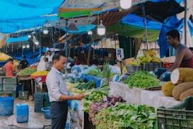 A bustling market scene with a variety of fresh produce on display, including bananas, greens, and jackfruits. Several people are engaged in buying and selling activities under bright blue tarps that serve as makeshift roofs. Crates of vegetables and herbs fill the stalls, and the atmosphere is lively with vendors attending to their goods.