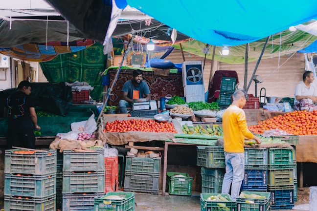 A bustling fruit and vegetable market stall with a variety of fresh produce. Crates filled with tomatoes, cucumbers, and other vegetables are neatly stacked. There are vendors interacting with customers and organizing the produce. A colorful tarp provides shelter, creating a vibrant atmosphere.