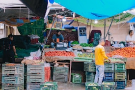 A bustling fruit and vegetable market stall with a variety of fresh produce. Crates filled with tomatoes, cucumbers, and other vegetables are neatly stacked. There are vendors interacting with customers and organizing the produce. A colorful tarp provides shelter, creating a vibrant atmosphere.