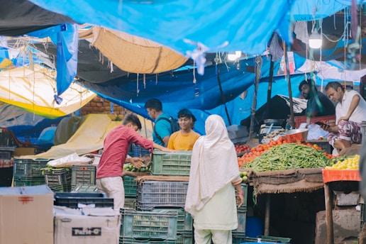 A busy marketplace scene with several people engaged in lively interactions around vegetable stalls. Various fresh produce including green beans, tomatoes, and yellow peppers are displayed prominently. The stalls are covered with colorful tarps providing shade. The atmosphere is bustling and vibrant.