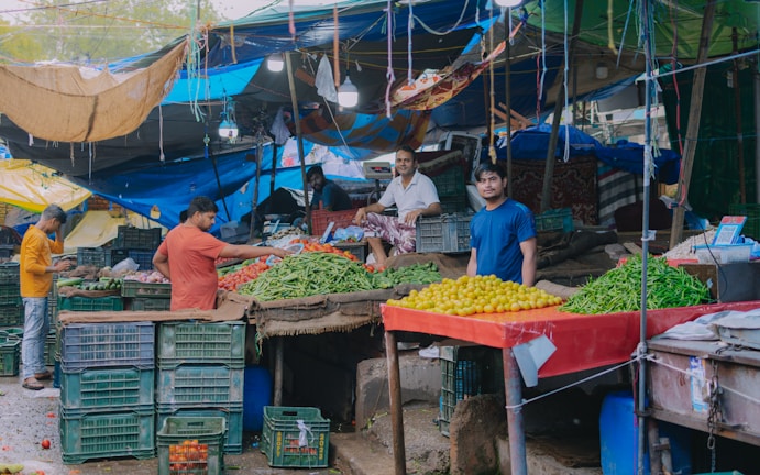 A bustling market scene with several vendors displaying fresh produce. Colorful awnings and tarps cover the stalls, providing shelter. Crates of vegetables such as green beans, yellow tomatoes, and others are prominently displayed. People interact around the stalls, with one vendor standing near a table filled with produce.