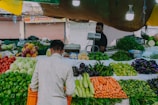 A market stall displaying a variety of fresh vegetables including carrots, cucumbers, bell peppers, and eggplants. A vendor stands behind the counter, with a customer in a white shirt picking vegetables. The setting is illuminated by hanging lights, and the backdrop includes closed shop shutters.