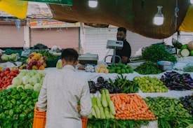 A market stall displaying a variety of fresh vegetables including carrots, cucumbers, bell peppers, and eggplants. A vendor stands behind the counter, with a customer in a white shirt picking vegetables. The setting is illuminated by hanging lights, and the backdrop includes closed shop shutters.