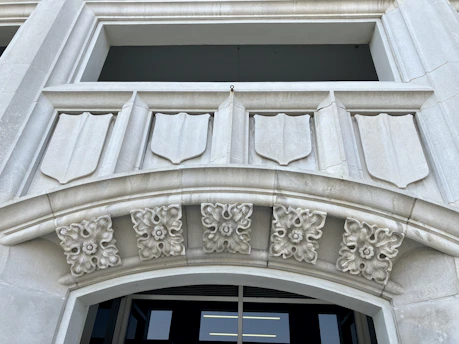 A detailed architectural design featuring a stone facade with shield-like decorative elements. There are intricate floral motifs adorning the archway, adding to the classical aesthetic. The structure appears to be made of light gray stone and the background shows an opening, possibly a window or entrance.