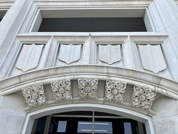 A detailed architectural design featuring a stone facade with shield-like decorative elements. There are intricate floral motifs adorning the archway, adding to the classical aesthetic. The structure appears to be made of light gray stone and the background shows an opening, possibly a window or entrance.