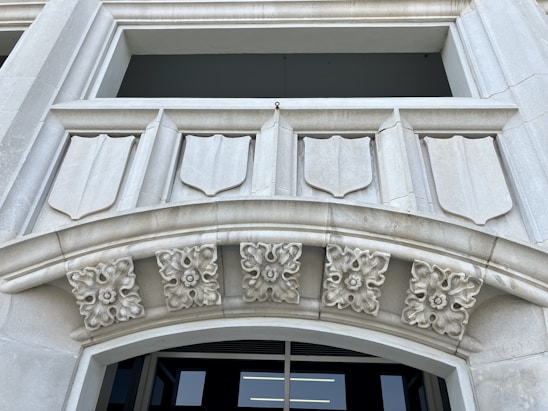 A detailed architectural design featuring a stone facade with shield-like decorative elements. There are intricate floral motifs adorning the archway, adding to the classical aesthetic. The structure appears to be made of light gray stone and the background shows an opening, possibly a window or entrance.