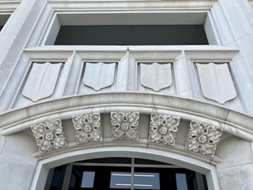 A detailed architectural design featuring a stone facade with shield-like decorative elements. There are intricate floral motifs adorning the archway, adding to the classical aesthetic. The structure appears to be made of light gray stone and the background shows an opening, possibly a window or entrance.