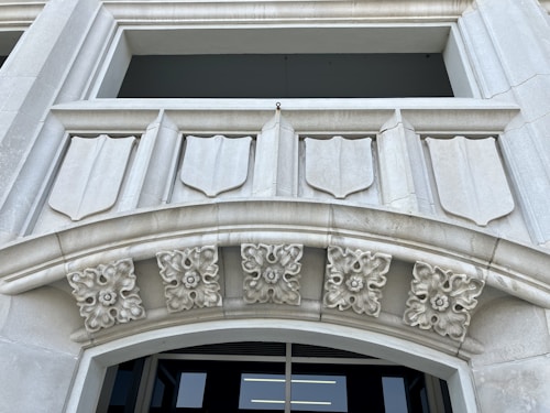 A detailed architectural design featuring a stone facade with shield-like decorative elements. There are intricate floral motifs adorning the archway, adding to the classical aesthetic. The structure appears to be made of light gray stone and the background shows an opening, possibly a window or entrance.