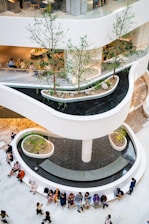 A multi-level indoor space with modern architectural design elements, featuring a series of curved walkways and planters with small trees. Water cascades down a central column surrounded by a large circular seating area. People are seated and standing around the space, engaging in various activities such as talking and using smartphones.