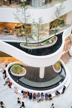 A multi-level indoor space with modern architectural design elements, featuring a series of curved walkways and planters with small trees. Water cascades down a central column surrounded by a large circular seating area. People are seated and standing around the space, engaging in various activities such as talking and using smartphones.