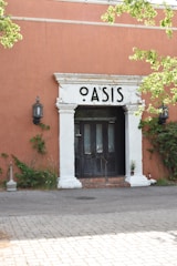 A building entrance marked with a sign reading 'Oasis' above dark wooden doors. There is a brick step leading up to the doors, and the surrounding wall is painted a warm terracotta color. Lush green ivy and plants crawl up the walls on either side of the entrance. A lantern-style light fixture is mounted on the wall to the left of the doors. The foreground features a cobblestone pathway.