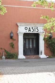 A building entrance marked with a sign reading 'Oasis' above dark wooden doors. There is a brick step leading up to the doors, and the surrounding wall is painted a warm terracotta color. Lush green ivy and plants crawl up the walls on either side of the entrance. A lantern-style light fixture is mounted on the wall to the left of the doors. The foreground features a cobblestone pathway.