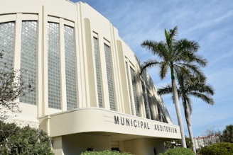 A large municipal auditorium with art deco architecture features tall, vertical glass block windows and rounded edges. In front of the building, a few palm trees are visible under a blue sky. The area is surrounded by some greenery.