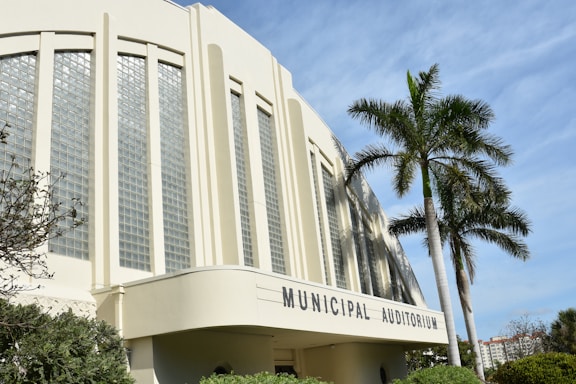 A large municipal auditorium with art deco architecture features tall, vertical glass block windows and rounded edges. In front of the building, a few palm trees are visible under a blue sky. The area is surrounded by some greenery.