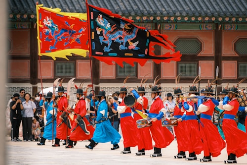 A group of individuals dressed in traditional Korean military attire, performing a ceremonial march in front of a historic building. The uniforms are vibrant red with blue belts, and the participants carry long spears and rectangular shields. Two large, colorful flags depicting dragons are prominently displayed above the marchers. Spectators are gathered in the background.