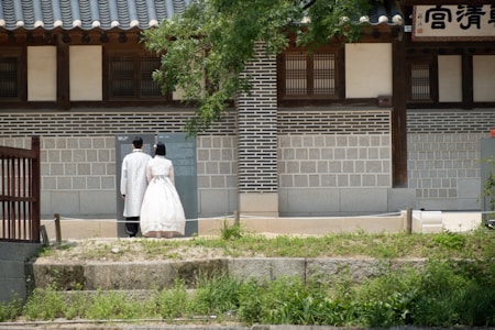 A couple dressed in traditional attire stands side by side, facing an information board outside a historical building. The structure features intricate brickwork and wooden elements, with greenery in the foreground.