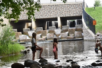 Children are playing near a water structure that consists of multiple levels with cascading waterfalls, made with a stone facade. Large rocks and shallow water surround the children as they stand and wade. A vibrant green lawn and trees frame the scene, with a fence and sign visible on the side.