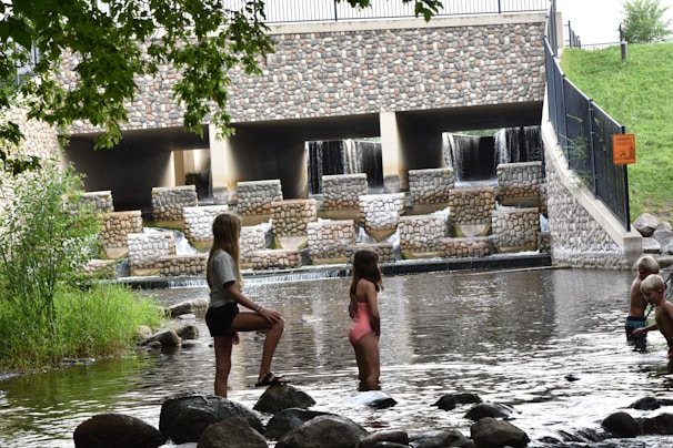 Children are playing near a water structure that consists of multiple levels with cascading waterfalls, made with a stone facade. Large rocks and shallow water surround the children as they stand and wade. A vibrant green lawn and trees frame the scene, with a fence and sign visible on the side.