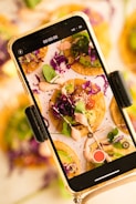 A smartphone is capturing an image of colorful, garnish-topped mini tacos on a plate. The tacos are adorned with vibrant purple cabbage, leafy greens, sliced avocado, and a drizzle of creamy sauce. The screen shows a close-up of the dish, revealing details of the toppings and textures.