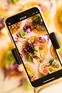 A smartphone is capturing an image of colorful, garnish-topped mini tacos on a plate. The tacos are adorned with vibrant purple cabbage, leafy greens, sliced avocado, and a drizzle of creamy sauce. The screen shows a close-up of the dish, revealing details of the toppings and textures.