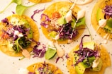 A collection of vibrant tacos topped with avocado slices, shredded purple cabbage, greens, and drizzles of a creamy sauce. Each taco is garnished with sesame seeds, resting on a light-colored surface.