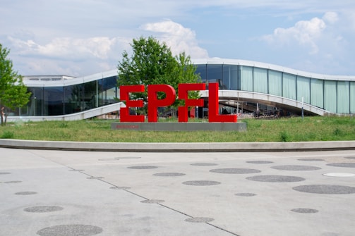 Large red letters spelling out 'EPFL' stand in front of a modern, curved glass building. The surrounding area features green grass and a few trees. The sky is partly cloudy, creating a bright and open atmosphere.