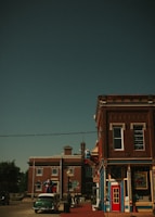 A vintage-style street scene features brick buildings with ornate trim. Statues resembling iconic superheroes are displayed on the building and street, adding a whimsical touch. A classic car is parked in front, enhancing the retro atmosphere. The sky is clear and the overall scene evokes a sense of nostalgia.