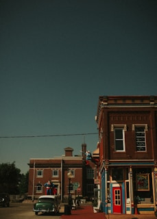 A vintage-style street scene features brick buildings with ornate trim. Statues resembling iconic superheroes are displayed on the building and street, adding a whimsical touch. A classic car is parked in front, enhancing the retro atmosphere. The sky is clear and the overall scene evokes a sense of nostalgia.